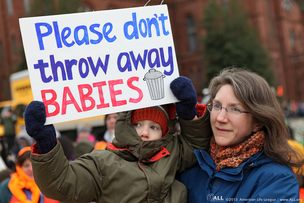 Child holds a sign that reads "Please don't throw away babies"